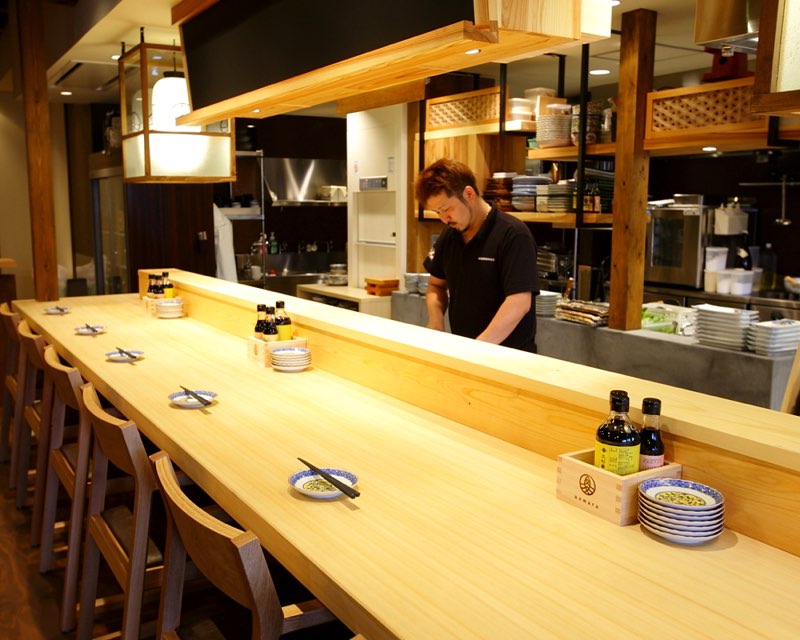 白木の一枚板カウンター越しに板前が仕込みをする、魚丸 彦根店のカウンター席とオープンキッチン