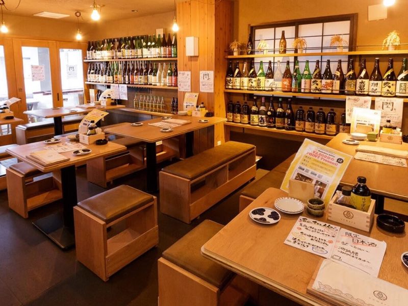 日本酒の一升瓶が並ぶ棚と1階のテーブル席（炉端と日本酒 魚丸 守山店）