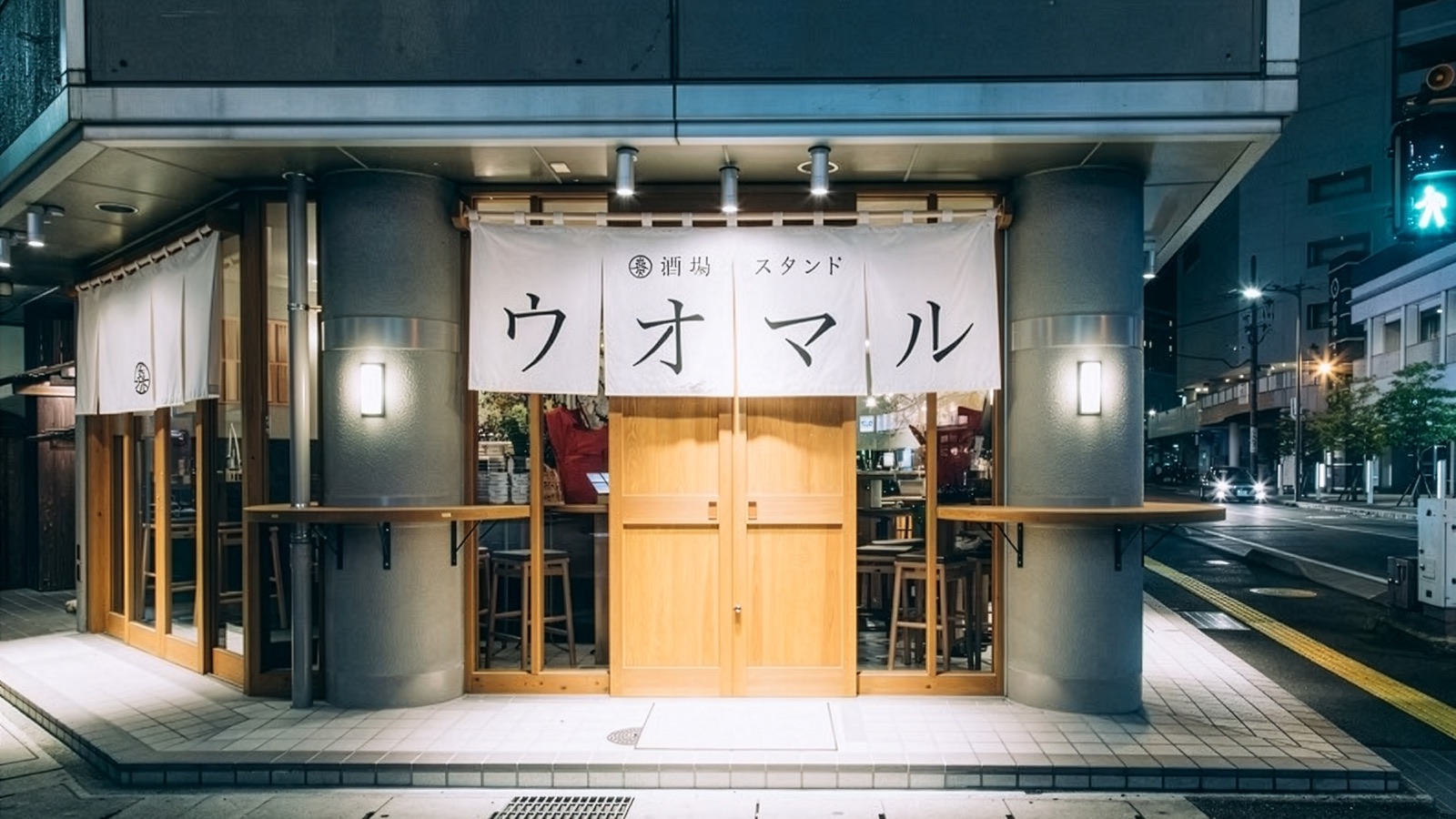 夜の「酒場スタンド ウオマル」の外観。木の引き戸の上に白い暖簾が下がり、店内のカウンターに明かりが灯っている