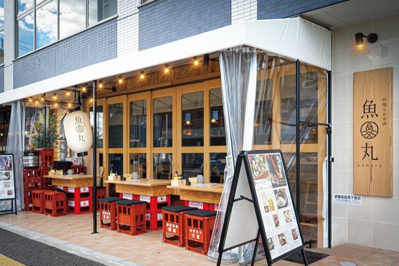 「炉端と日本酒 魚丸」の木看板と、赤いビールケースを椅子にしたテラス席が並ぶ魚丸 野洲店の外観